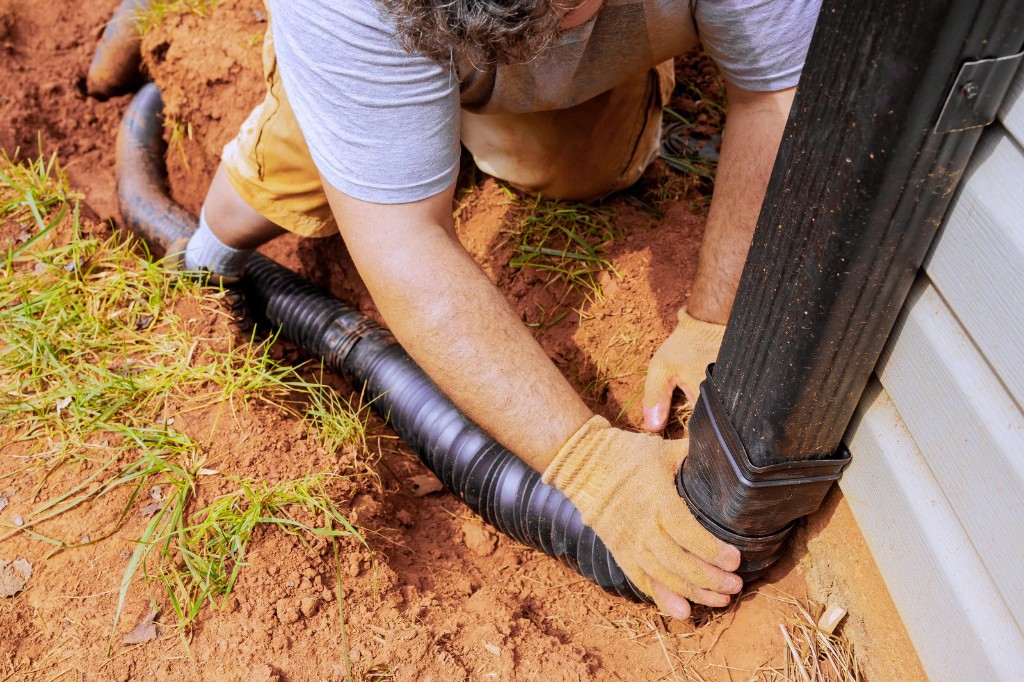 Worker in gloves connecting a corrugated drainage pipe to a downspout at the base of a house