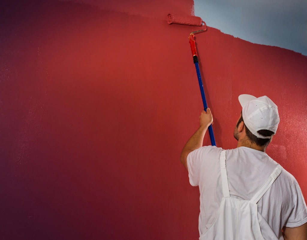 Painter in white overalls rolling red paint onto an interior wall with an extension pole